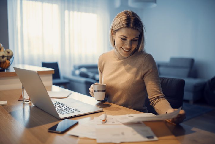 Mujer sonriendo con una taza de café revisando los documentos financieros de su negocio, considerando la posibilidad de refinanciación.