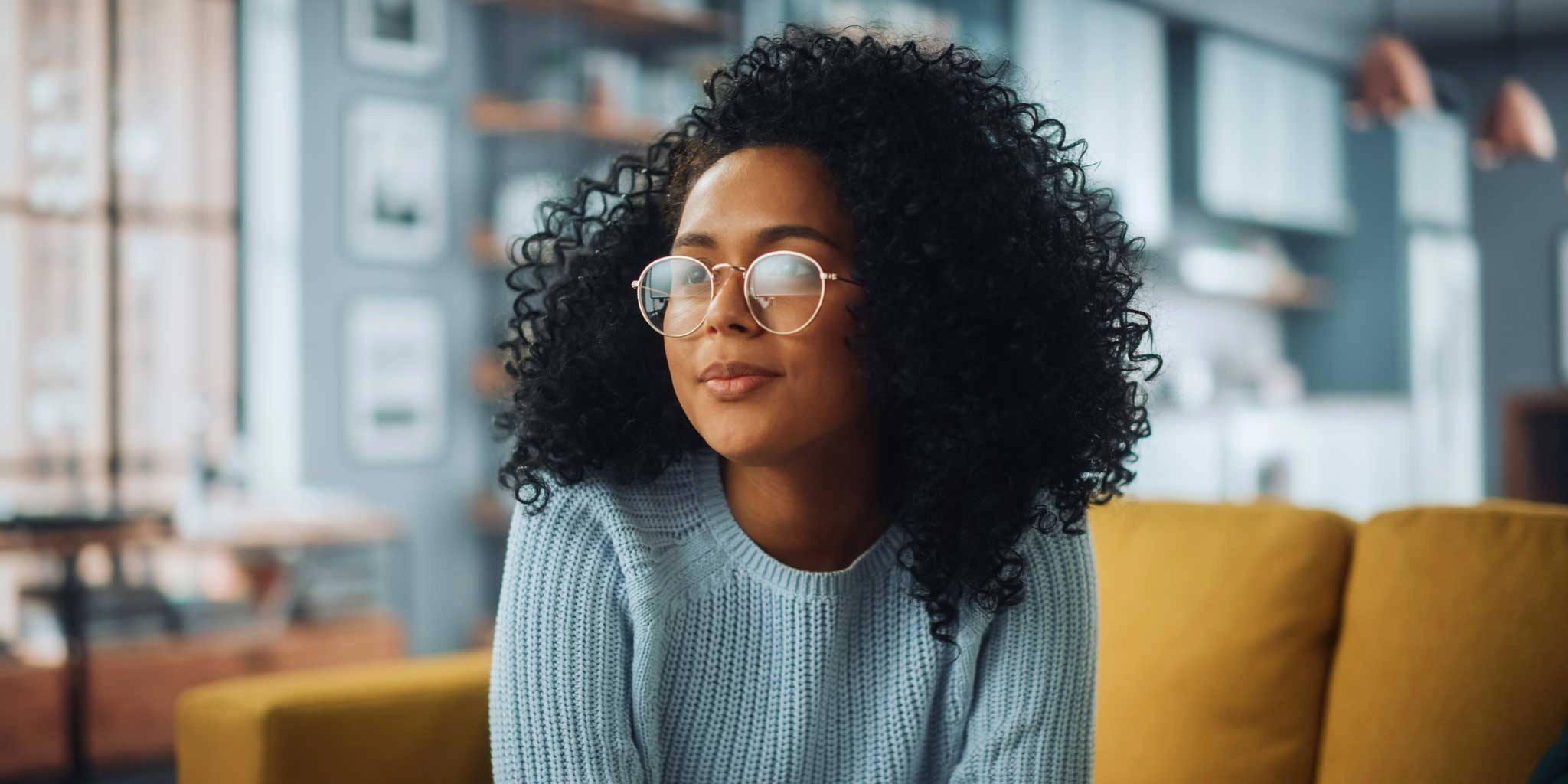 Portrait of a Beautiful Authentic Latina Female with Afro Hair Wearing Light Blue Jumper and Glasses. She Looks Away and Thinking about Life. Successful Woman Resting in Bright Living Room.