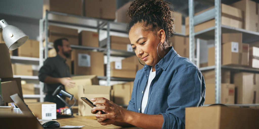 Inventory Manager Using Smartphone to Scan a Barcode on Parcel, Preparing a Small Cardboard Box for Postage. Black Multiethnic Small Business Owner Working on Laptop in Warehouse.