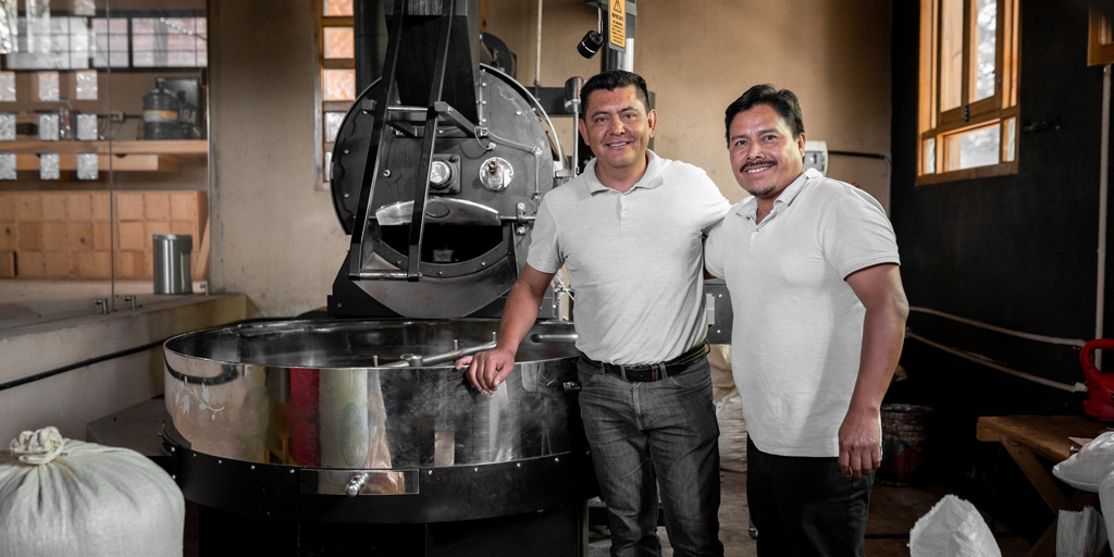 Two Hispanic men are smiling next to their big coffee roaster machine