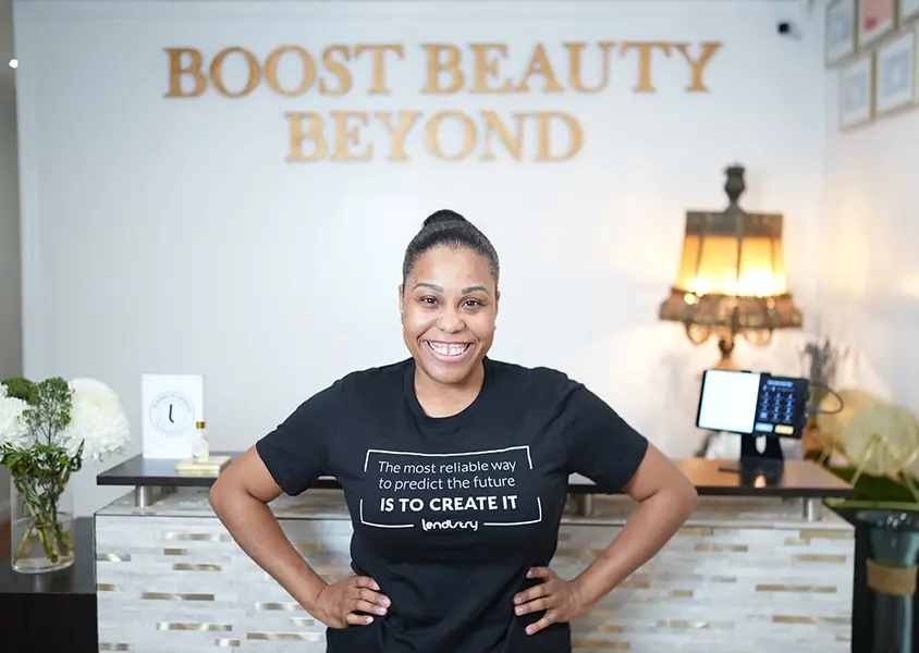 Betzayda David of Boost Beauty Beyond (BBB) posing with hands at the hips smiling at the camera standing at the reception area in front of store sign